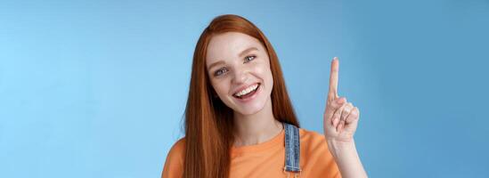 Lively active energized happy smiling hepful redhead female coworker showing you awesome promo pointing index finger up grinning introduce cool product recommend try out blue background photo