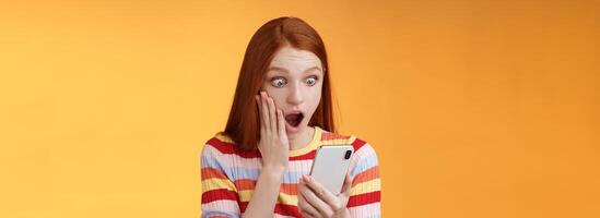 Omg what heck. Portrait shocked concerned young redhead sensitive impressed redhead woman stare smartphone display touch cheek drop jaw stunned surprised standing orange background hold phone photo