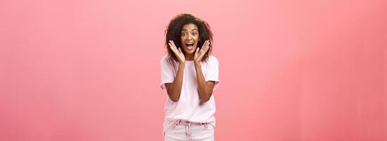 Indoor shot of expressive excited attractive dark-skinned adult woman in casual outfit yelling from amazement and joy holding palms near face reacting on awesome news with excitement and happiness photo