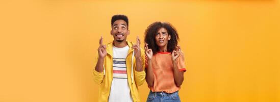 Couple of friends praying and looking up in sky intense crossing fingers for good luck clenching teeth faithfully hoping parents not saw them smoking standing together over orange background photo