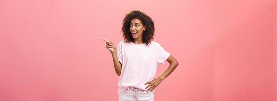 Hey wanna go there. Portrait of intrigued pleased good-looking stylish african woman on vacation wearing summer clothes pointing and gazing right with curious look over pink background photo