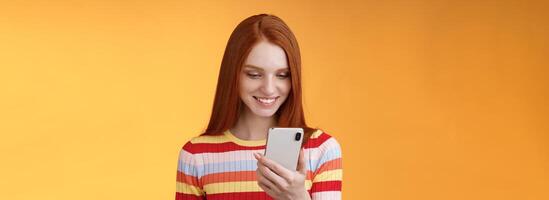 Charming modern redhead girl college student checking message box holding smartphone look happy smiling delighted cellphone display receive hundred likes photo post online, orange background