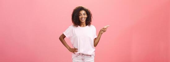 Friendly chill and relaxed attractive dark-skinned girlfriend with curly hairstyle holding hand on hip and pointing left with assured pleased expression showing where she bought cool outfit photo