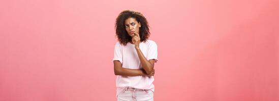 Suspicious displeased dark-skinned young curly-haired woman in trendy t-shirt and shorts saying hmm touching chin with finger frowning and staring left doubtful and unsure having concern photo