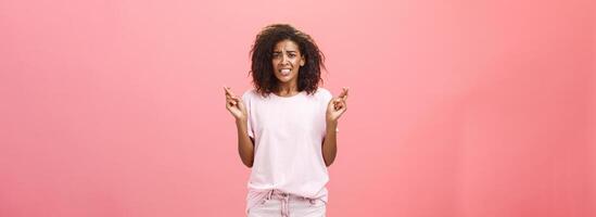 Hope she gets positive result. Portrait of worried intense hopeful good-looking young african american female coworker frowning clenching teeth and crossing fingers for good luck while making wish photo