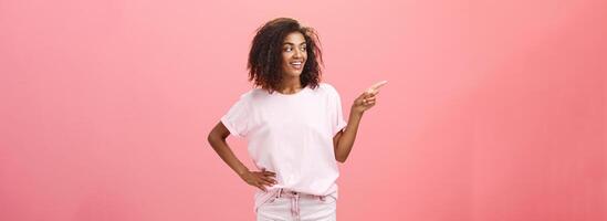 Girl checking out nice body of guy passing by. Charming confident dark-skinned female with curly hairstyle holding hand on hip looking and pointing left discussing cool copy space over pink wall photo