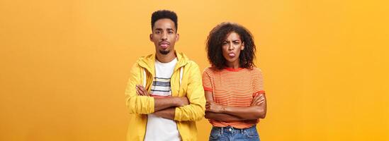 Indoor shot of african american siblings being displeased and annoyed showing bad tempber behaving childish sticking out tongue standing together with crossed hands on chest over orange background photo