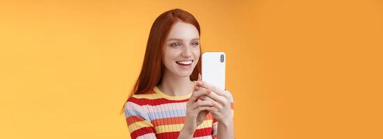 Redhead girl having fun recording hilarious friend actions hold smartphone look display amused shooting funny video telephone standing orange background satisfied smiling delighted photo