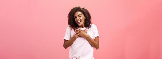 Portrait of charming delighted african american woman with curly haircut holding palms on heart pleased and grateful thanking friend for help smiling happily and thankful at camera over pink wall photo