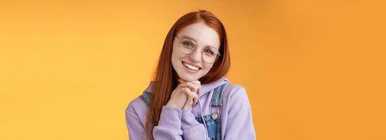 Tender romantic lovely redhead girlfriend leaning palms tilting head cute flirty giggling smiling camera white teeth wearing glasses hoodie overalls near orange background, sensually laughing photo