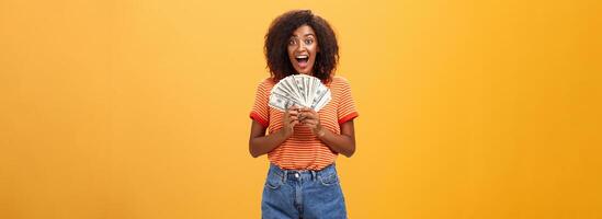 Woman cannot hide happiness winning lottery. Portrait of surprised and happy lucky african american young woman with curly hair holding bunch of money and yelling from delight receiving paycheck photo