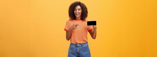 Girl showing picture in phone bragging about her cool vacation. Charming friendly and sociable african american woman with curly hair holding cellphone pointing at smartphone screen over orange wall photo