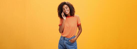 Stylish dark-skinned girl making casual phone call to friend telling all details of after romantic date standing pleased and carefree over orange background in striped t-shirt gazing left with grin photo