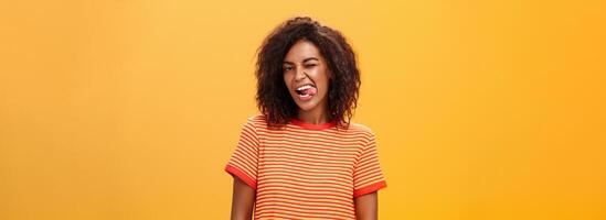 Portrait of daring and emotive confident flirty woman with afro hairstyle winking joyfully showing tongue posing carefree and enthusiastic against orange background flirting with hot guy photo