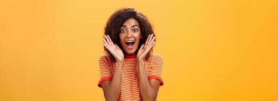 Waist-up shot of impressed excited talkative dark-skinned woman with curly hair and perfect skin yelling from happiness and amazement shaking palms near face cheering for awesome sale over orange wall photo