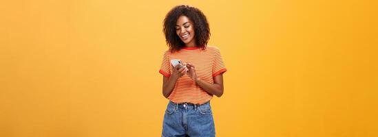 Stylish carefree girl texting friend come over standing pleased over orange wall in stylish denim shorts typing message or scrolling news in internet via smartphone gazing at device screen with smile photo