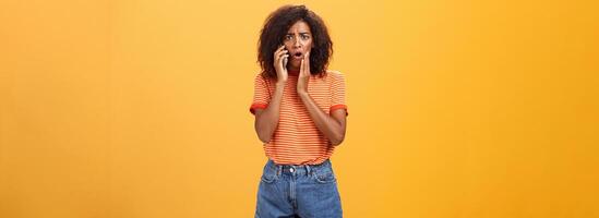 Woman receiving disturbing call feeling emapthy and sorry for poor friend getting in trouble holding cellphone near ear touching cheek concerned frowning standing sad, worried over orange background photo