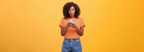 Indoor shot of shocked stunned african american young woman with afro hairstyle staring surprised and excited at smartphone screen holding cellphone reading amazing message over orange wall photo