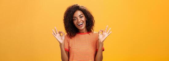 Waist-up shot of chill outgoing calm girlfriend with confident look in trendy striped t-shirt showing okay or excellent gestur and smiling broadly assuring everything alright over orange background photo