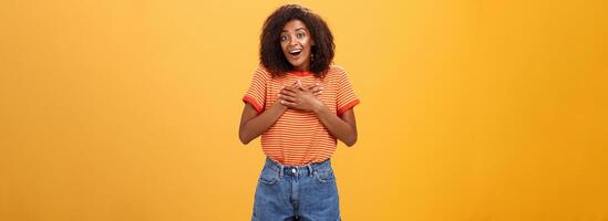 Cute delighted african american woman being grateful and thankful for help standing pleased over orange wall holding palms on breast thanking friend for rescuing grinning joyfully at camera photo