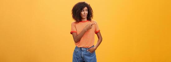Girl looking at doubtful unimpressive perfomance of model. Portrait of displeased confused good-looking dark-skinned female with afro hairstyle looking and pointing left with scorn and indifference photo