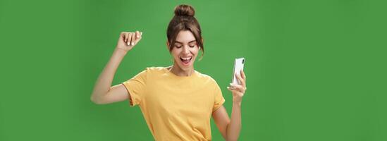 Cool and stylish happy woman with tattoo and gap teeth in yellow t-shirt dancing joyfully listening music in wireless earphones holding smartphone closing eyes and smiling from delight over green wall photo