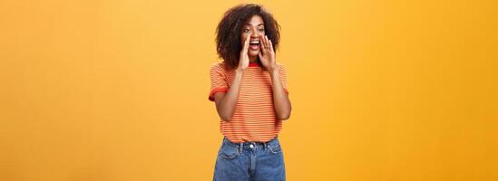 Girl calling coworker help out turning left with carefree joyful look holding hands near opened mouth to shout louder asking friend come closer while yelling on distance over orange background photo