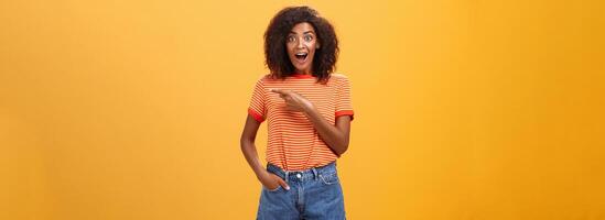 Hey awesme copy space there. Portrait of impressed and surprised enthusiastic young african american female with afro hairstyle pointing right astonished looking energized and curious over orange wall photo