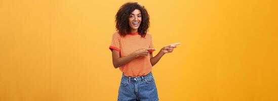 Ask him. Portrait of friendly and joyful good-looking stylish female shop assistant with curly hair and dark skin pointing left with both hands, smiling assured and entertained over orange background photo