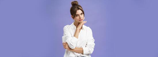 Portrait of tender and feminine stylish woman in white blouse posing sensually and flirty touching chin gazing daring at camera posing against purple background with self-assured expression photo