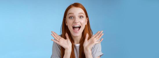 Happy cheerful rejoicing young pretty redhead supportive girlfriend clasp hands joyfully yelling congratulations raising hands amused wide eyes surprised getting awesome good news, blue background photo