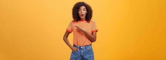 estudio Disparo de elegante ajuste hembra modelo en mezclilla pantalones cortos y camiseta jadeando impresionado señalando Derecha mientras preguntando pregunta siendo interesado durante observando clase maestra terminado naranja antecedentes foto