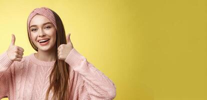 Totally approve. Girl agrees taking part in interesting activity smiling joyful, staying positive showing thumbs up in support, recommend super cool service. posing delighted against yellow wall photo