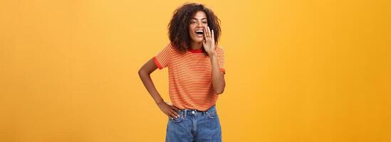 Girl calling sister come to phone yelling out loud holding palm near opened mouth to be scream louder so friend can hear holding arm on waist gazing left with carefree pleased look over orange wall photo