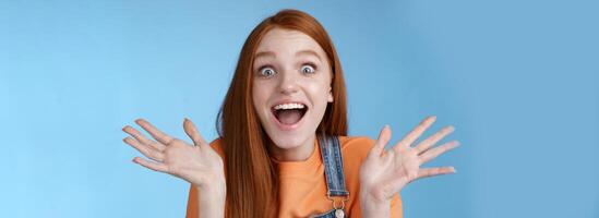 Amused speechless surprised astonished redhead girl hear exciting positive news smiling gladly wide eyes thrilled raised hand triumphing win lottery celebrating victory success, blue background photo