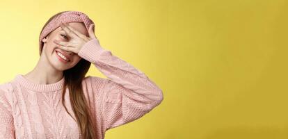 Blushing cute girl feeling positive, smiling happy joyful covering eyes with palm peeking through fingers delighted, playful, tilting head tender and lovely gazing at camera against yellow background photo