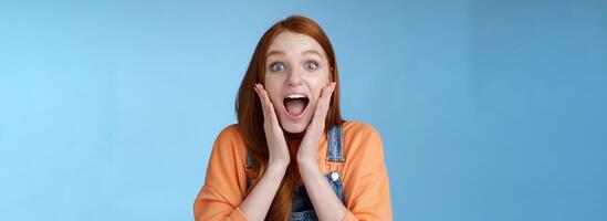 Excited thrilled young emotional enthusiasitc ginger girl teenage college student yelling amused smiling broadly receive positive good news look surprised camera touch face astonished blue background photo