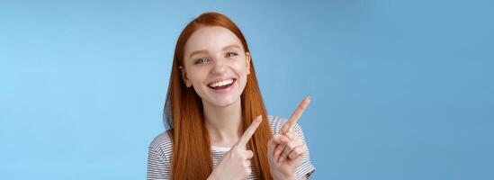 Happy carefree smiling redhead caucasian girl ginger straight hairstyle pointing upper left corner laughing joyfully amused cool promo giving recommendation try new product, standing blue background photo