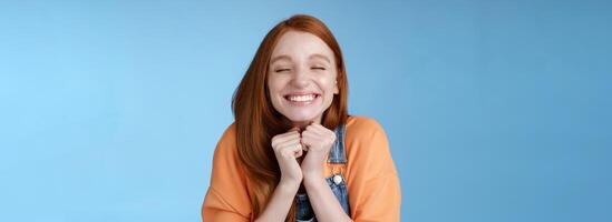 Eager rejoicing thrilled pretty young redhead girl close eyes dreamy smiling receive great result scholarchip triumphing joyfully grinning squeez hands excited, standing blue background very happy photo