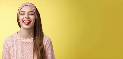 Positive entertained cute funny glamour young european girl in sweater, knitted trendy headband smiling fooling around showing tongue playfully, mocking friend enjoying sunny day over yellow wall photo