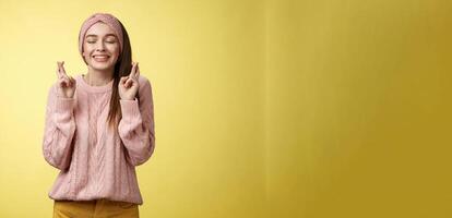 Girl wishing wellness wanting dream come true, cross fingers dreamy, close eyes waiting miracle, anticipating good news, posing excited and joyful against yellow background in knitted warm sweater photo