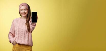 mejor teléfono inteligente aquí usted ir. encantador saliente joven bonito mujer en de punto suéter, venda extensión mano con teléfono demostración artilugio pantalla sonriente recomendando Teléfono móvil terminado amarillo pared foto