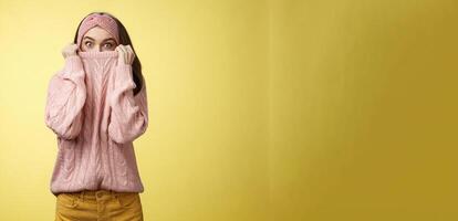 Girl hiding in collar wearing knitted sweater over nose popping eyes, staring bugged eyes at camera, hiding face reacting amazed and shocked, feeling scared, standing in stupor over yellow background photo