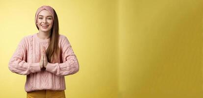 Please do me favour. Charming young female student wearing knitted sweater, headband pressing palms together in pray begging, asking for help smiling thankful, grateful for effort over yellow wall photo