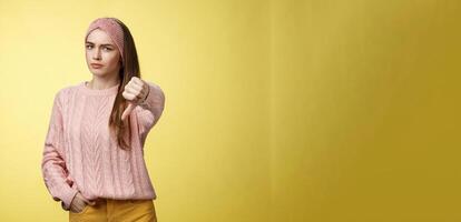 Not cool. Frustrated displeased and disagree cute young woman wearing knitted sweater frowning showing thumb-down smirking judgemental, expressing disapproval of unwell situation over yellow wall photo