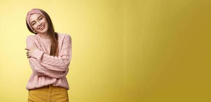 Cozy charming friendly young smiling girl in knitted warm comfortable sweater, grinning joyfully tilt head emracing herself, crossing arms, hugging feeling safe and happy, posing against yellow wall photo