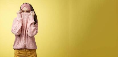 Sad silly, gloomy cute young girl hiding face in sweater collar pulling clothes on nose frowning displeased, unhappy, looking indignant and reluctant standing disappointed over yellow background photo