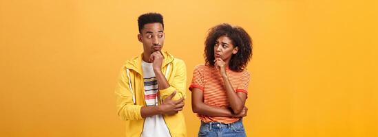 Silly confused girl standing gloomy and worried pursing lips and frowning holding hand on chin looking at boyfriend confused while guy glancing at girlfriend with curious expression over orange wall photo