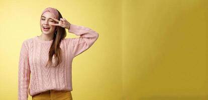 Cheerful happy glamour young european woman in pink knitted sweater, wearing headband, winking flirty and cute, showing victory or pease sign over eye, feeling excited and joyful over yellow wall photo