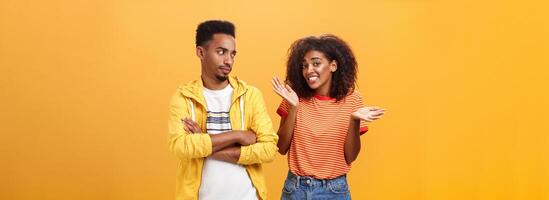 Guy thinks his friend weirdo making dumb thinks looking at cute girl with suspicious look crossing arms on chest raising eyebrow questioned while girlfriend saying sorry shrugging over orange wall photo